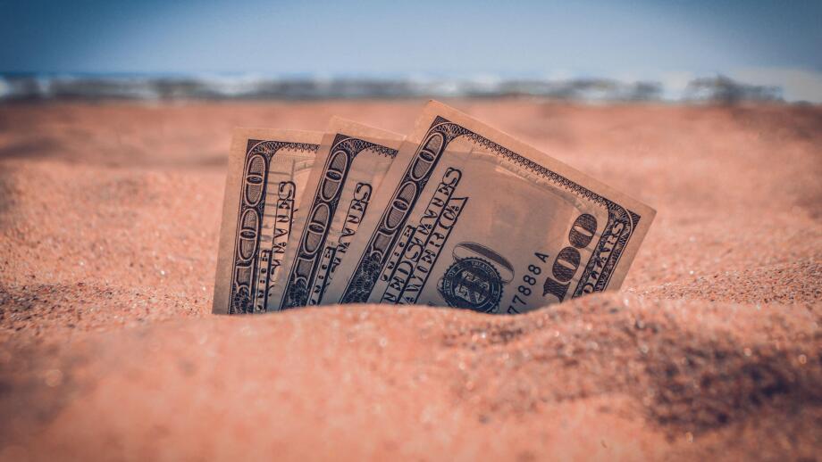 Money dolars half covered with sand lie on sandy beach near sea ocean waves on sunny summer day close-up. Money grows out of the ground. Concept finance money holiday relax vacation.