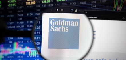 Goldman Sachs company logo on a website with blurry stock market developments in the background, seen on a computer screen through a magnifying glass