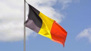 The national flag of Belgium flies up a flagpole on a beautiful summer day with a background of blue sky and white clouds.