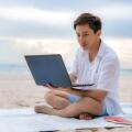 Young asian man working outdoor during his vacation time by laptop while sitting on the beautiful beach. Summer, holidays, vacation and happy people i