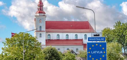 European Union border sign welcoming travelers to Latvia in Skaistkalne, with a historic church featuring a red roof and white walls in the background