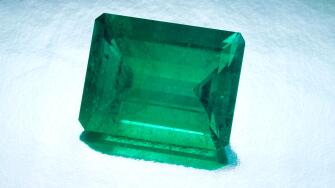 A large natural emerald cut emerald sits on a silver reflective background.