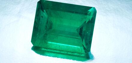 A large natural emerald cut emerald sits on a silver reflective background.