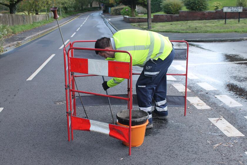 United utilities worker with hi vi jacket digging a hole in the road to locate source of water leak with safety barrier around him