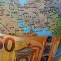 A fifty euro bill placed over the Strait of Hormuz on a world globe, symbolizing the impact of Middle East geopolitical tensions on European fuel pric
