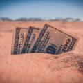 Money dolars half covered with sand lie on sandy beach near sea ocean waves on sunny summer day close-up. Money grows out of the ground. Concept finance money holiday relax vacation.