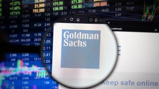 Goldman Sachs company logo on a website with blurry stock market developments in the background, seen on a computer screen through a magnifying glass