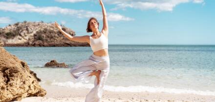 Woman with short brown hair wearing white sportswear practicing Tree Pose (Vrksasana) on a sandy beach beside the ocean with coastal rocks in the background.