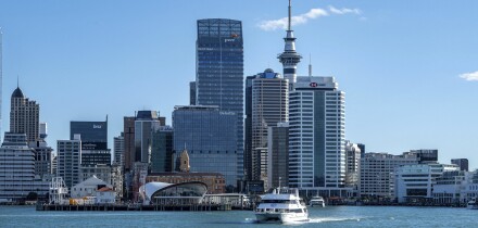 Modern urban skyline of central Auckland featuring high-rise commercial buildings, the iconic Sky Tower, and a passenger ferry crossing the harbor.