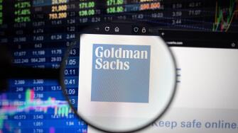 Goldman Sachs company logo on a website with blurry stock market developments in the background, seen on a computer screen through a magnifying glass