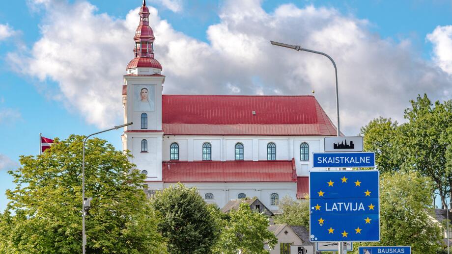 European Union border sign welcoming travelers to Latvia in Skaistkalne, with a historic church featuring a red roof and white walls in the background