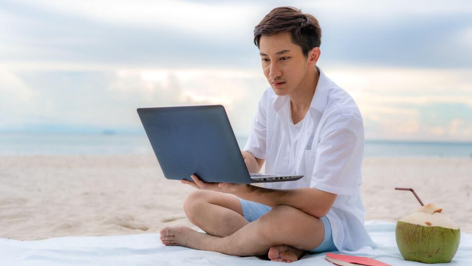 Young asian man working outdoor during his vacation time by laptop while sitting on the beautiful beach. Summer, holidays, vacation and happy people i