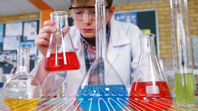 Examining red liquid in conical flask, boy wearing lab coat and goggles at school lab