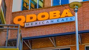 Qdoba Mexican Eats restaurant's exterior facade brand and logo signage in orange letters on red brick below reflective windows with lamppost.