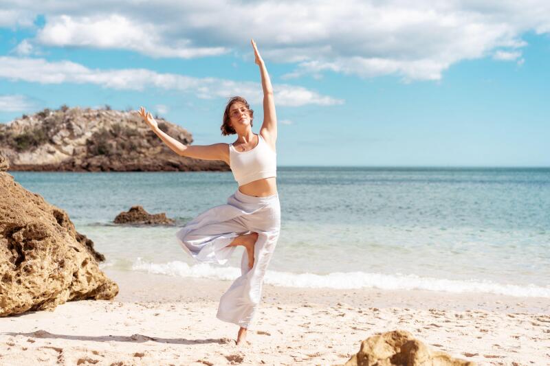 Woman with short brown hair wearing white sportswear practicing Tree Pose (Vrksasana) on a sandy beach beside the ocean with coastal rocks in the background.