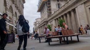 Pedestrians and seated office workers mingle in front of the historic neoclassical facade of the Bank of England on Threadneedle Street in the City of London, UK. Currently it is heavily covered in a massive, complex network of construction scaffolding. T