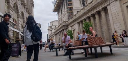 Pedestrians and seated office workers mingle in front of the historic neoclassical facade of the Bank of England on Threadneedle Street in the City of London, UK. Currently it is heavily covered in a massive, complex network of construction scaffolding. T