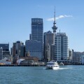 Modern urban skyline of central Auckland featuring high-rise commercial buildings, the iconic Sky Tower, and a passenger ferry crossing the harbor.