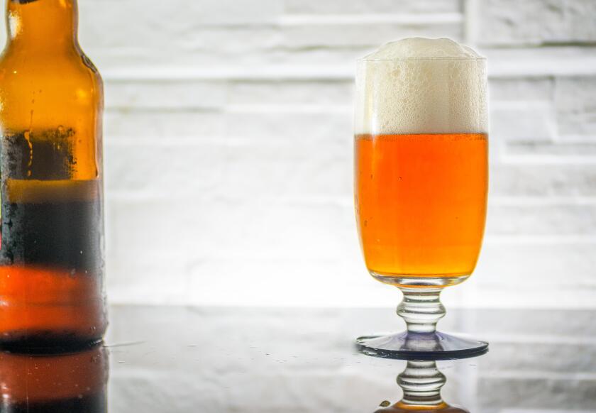 Beer glass full, with froth, and reflection, rugged stone wall as backdrop, and a beet bottle on the side, backlit