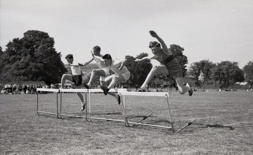 Secondary school sports day, England, c1960s, track & field, four schoolboys competing outside on.a grass track in a hurdles race, leaping over the barriers.