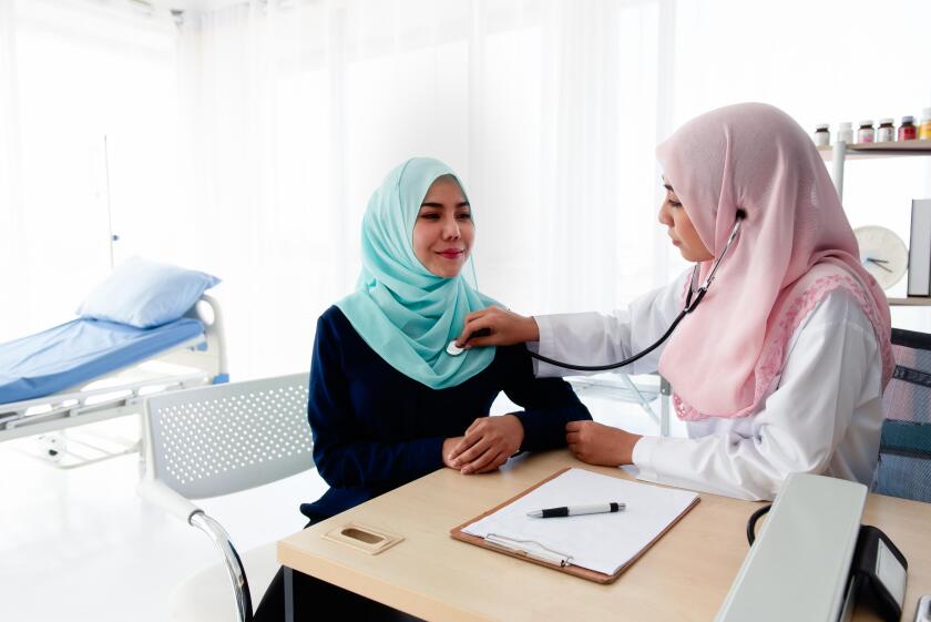 A young beautiful Muslim doctor is using a stethoscope to listen to the patient's heartbeat and write a report at the table in the hospital examinatio