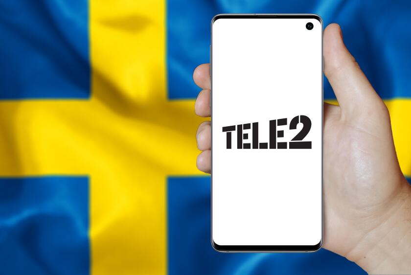 A man holds a smartphone displaying the logo of company Tele2 listed on OMX Stockholm. Swedish flag in the background. Credit: PIXDUCE