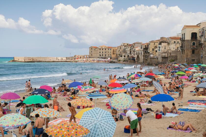 Cefalu, Sicily, Italy, crowds of people cover the sandy beach enjoying their holiday on a bright sunny August day with the old town buildings as a bac