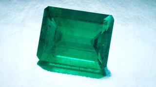 A large natural emerald cut emerald sits on a silver reflective background.