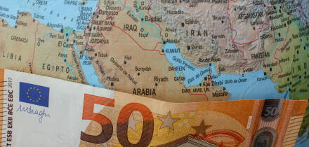 A fifty euro bill placed over the Strait of Hormuz on a world globe, symbolizing the impact of Middle East geopolitical tensions on European fuel pric
