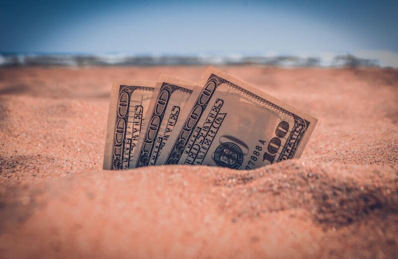 Money dolars half covered with sand lie on sandy beach near sea ocean waves on sunny summer day close-up. Money grows out of the ground. Concept finance money holiday relax vacation.