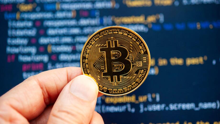 Hand holding a Bitcoin coin in front of computer code representing cryptocurrency development