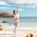Woman with short brown hair wearing white sportswear practicing Tree Pose (Vrksasana) on a sandy beach beside the ocean with coastal rocks in the background.