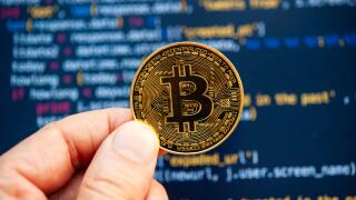 Hand holding a Bitcoin coin in front of computer code representing cryptocurrency development