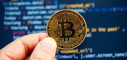 Hand holding a Bitcoin coin in front of computer code representing cryptocurrency development