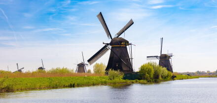 Panoramic view of multiple historic Dutch windmills lining the waterways in Kinderdijk, Netherlands, surrounded by green fields under a vast blue sky.