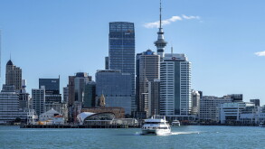 Modern urban skyline of central Auckland featuring high-rise commercial buildings, the iconic Sky Tower, and a passenger ferry crossing the harbor.