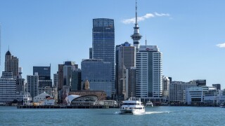 Modern urban skyline of central Auckland featuring high-rise commercial buildings, the iconic Sky Tower, and a passenger ferry crossing the harbor.