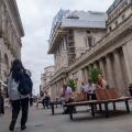 Pedestrians and seated office workers mingle in front of the historic neoclassical facade of the Bank of England on Threadneedle Street in the City of London, UK. Currently it is heavily covered in a massive, complex network of construction scaffolding. T
