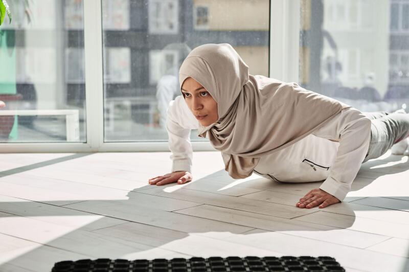 Determined Arab Muslim woman doing push-ups against the background of large windows at home on a beautiful sunny day. Home workout concept