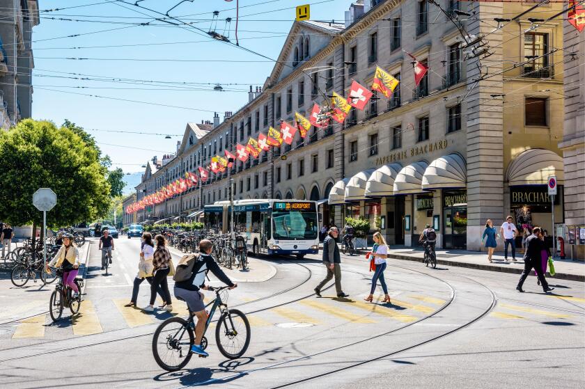 Pedestrians and cyclists are crossing the rue de la Corraterie in Geneva, with buildings decked with flags, as a bus is waiting at a red light.