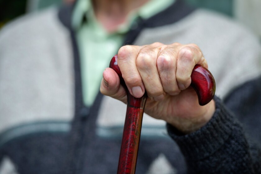An elderly old age pensioner sits with his hands on a walking stick in a care home.