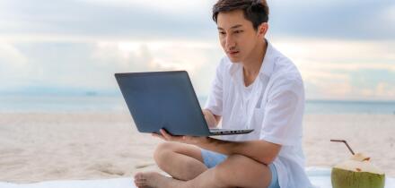Young asian man working outdoor during his vacation time by laptop while sitting on the beautiful beach. Summer, holidays, vacation and happy people i