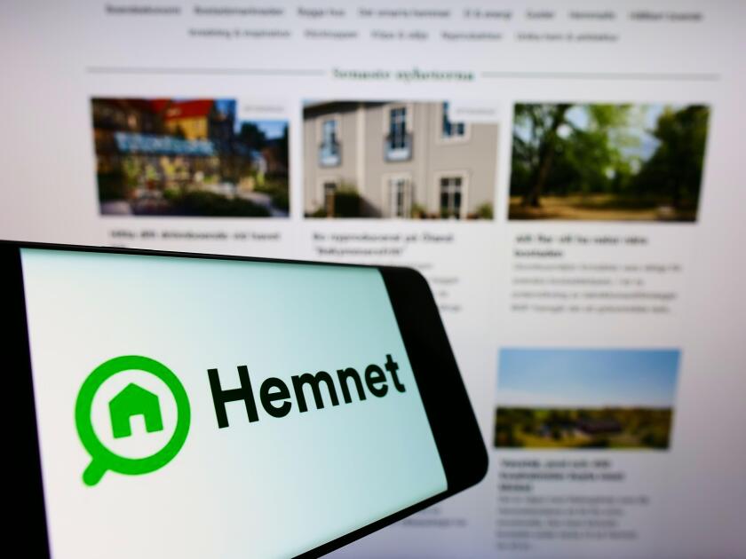 Mobile phone with logo of Swedish property platform company Hemnet AB on screen in front of business website. Focus on center-left of phone display.