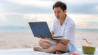 Young asian man working outdoor during his vacation time by laptop while sitting on the beautiful beach. Summer, holidays, vacation and happy people i