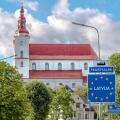European Union border sign welcoming travelers to Latvia in Skaistkalne, with a historic church featuring a red roof and white walls in the background