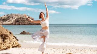 Woman with short brown hair wearing white sportswear practicing Tree Pose (Vrksasana) on a sandy beach beside the ocean with coastal rocks in the background.