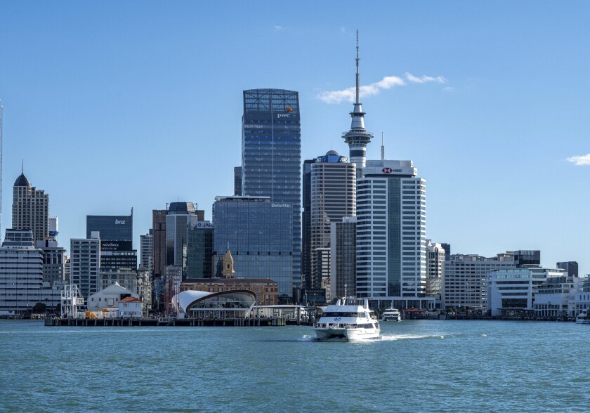 Modern urban skyline of central Auckland featuring high-rise commercial buildings, the iconic Sky Tower, and a passenger ferry crossing the harbor.