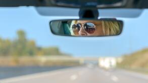Woman driving a car on a motorway with a view ahead through the windscreen and her glasses reflected in the rear view mirror