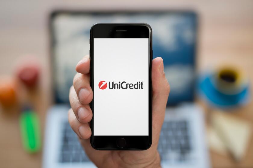 A man looks at his iPhone which displays the UniCredit logo, while sat at his computer desk (Editorial use only).