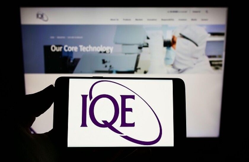 Person holding smartphone with logo of British semiconductor company IQE plc on screen in front of website. Focus on phone display.